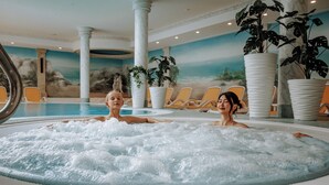 Un jacuzzi con dos personas, rodeado de sillones de descanso y grandes macetas, en una habitación con un mural de un paisaje.