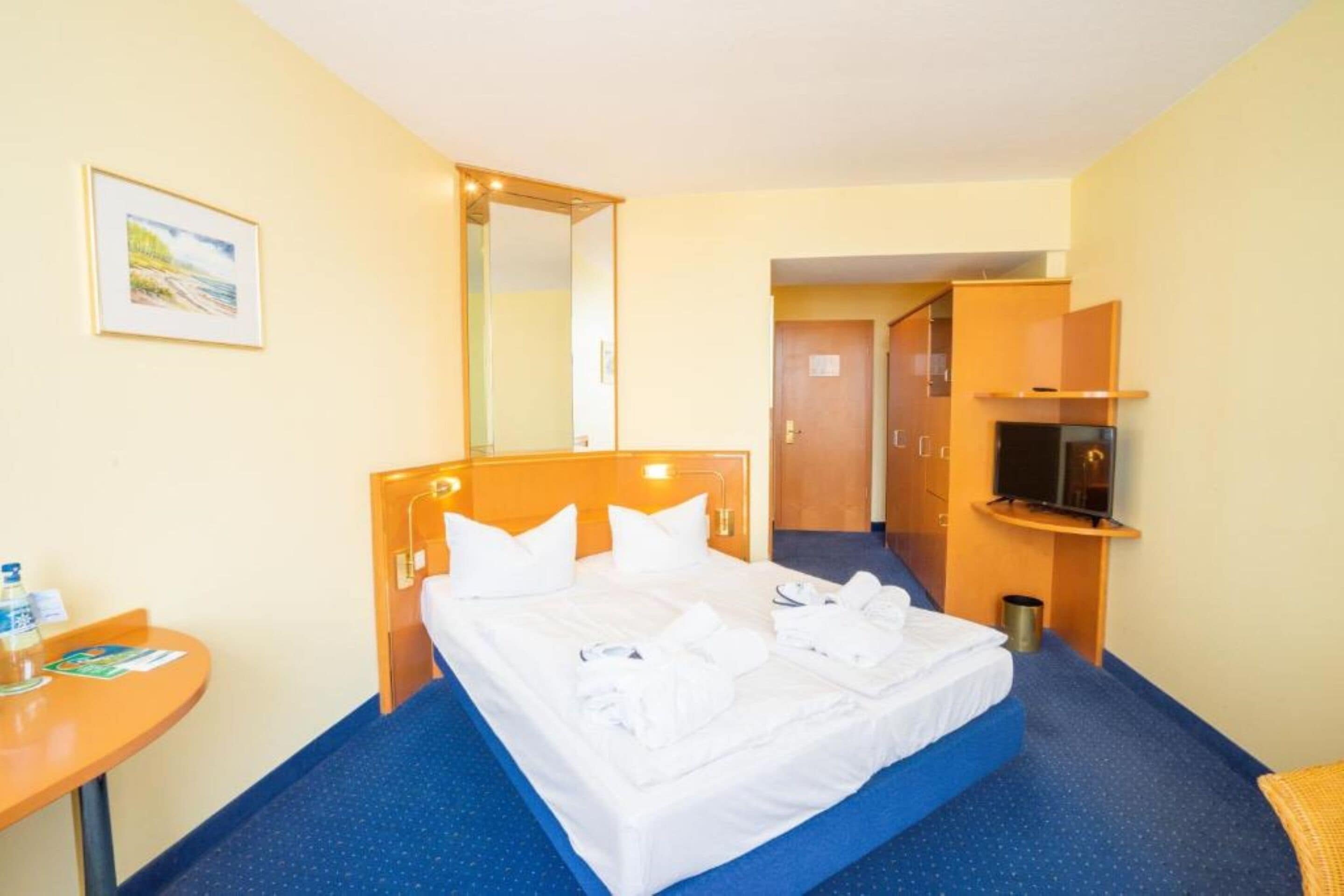 Une chambre d’hôtel avec un lit, une petite table sur laquelle se trouvent une bouteille et une brochure, un téléviseur posé sur un meuble et un miroir au mur.
