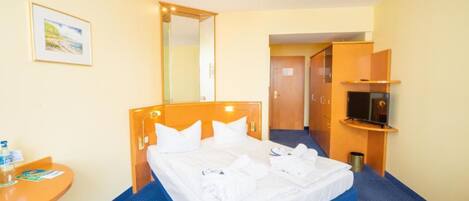 Une chambre d’hôtel avec un lit, une petite table sur laquelle se trouvent une bouteille et une brochure, un téléviseur posé sur un meuble et un miroir au mur.