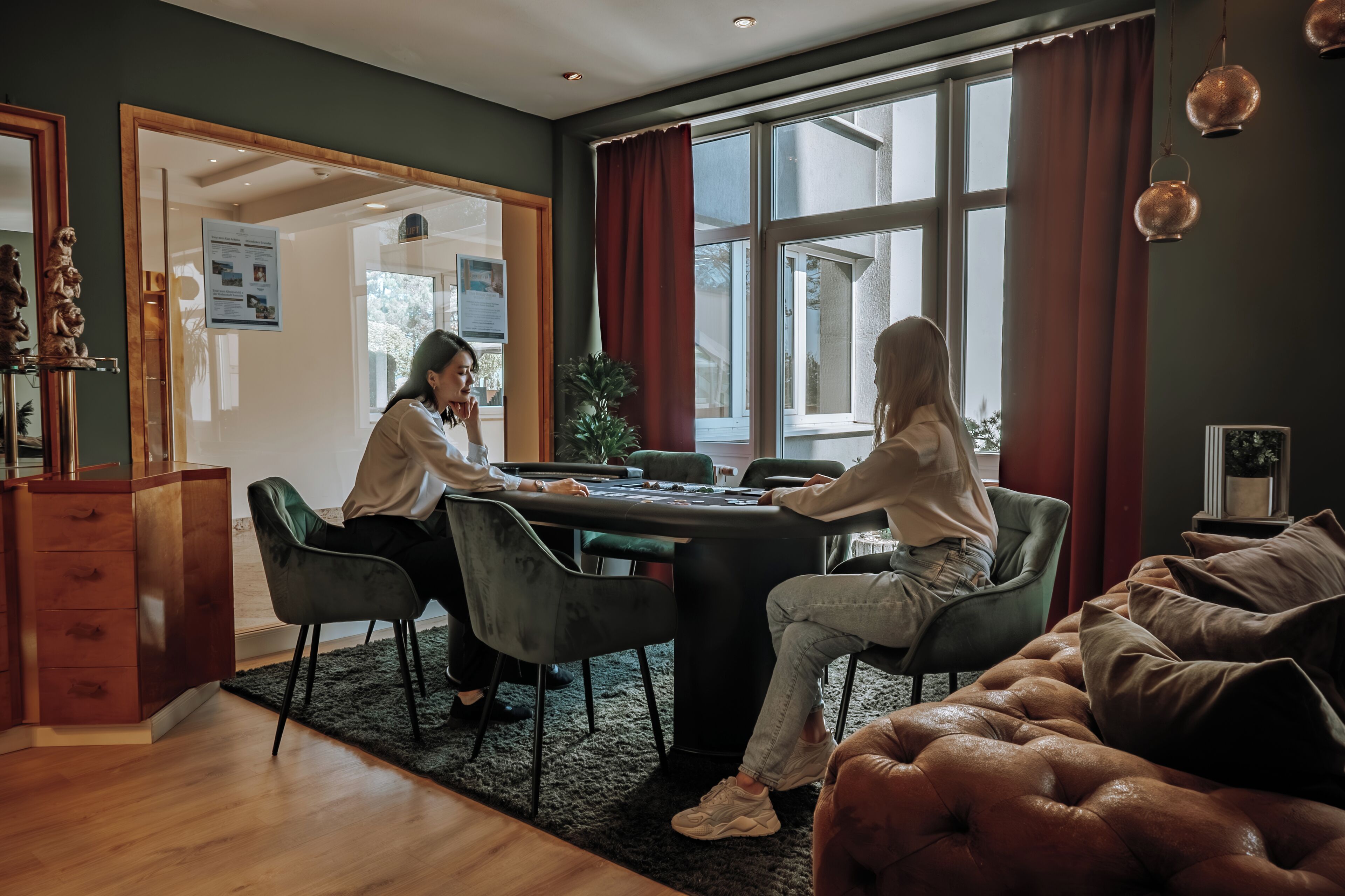 A modern living room with a wooden floor, a green sofa, a black gaming table, and a large window with red curtains.