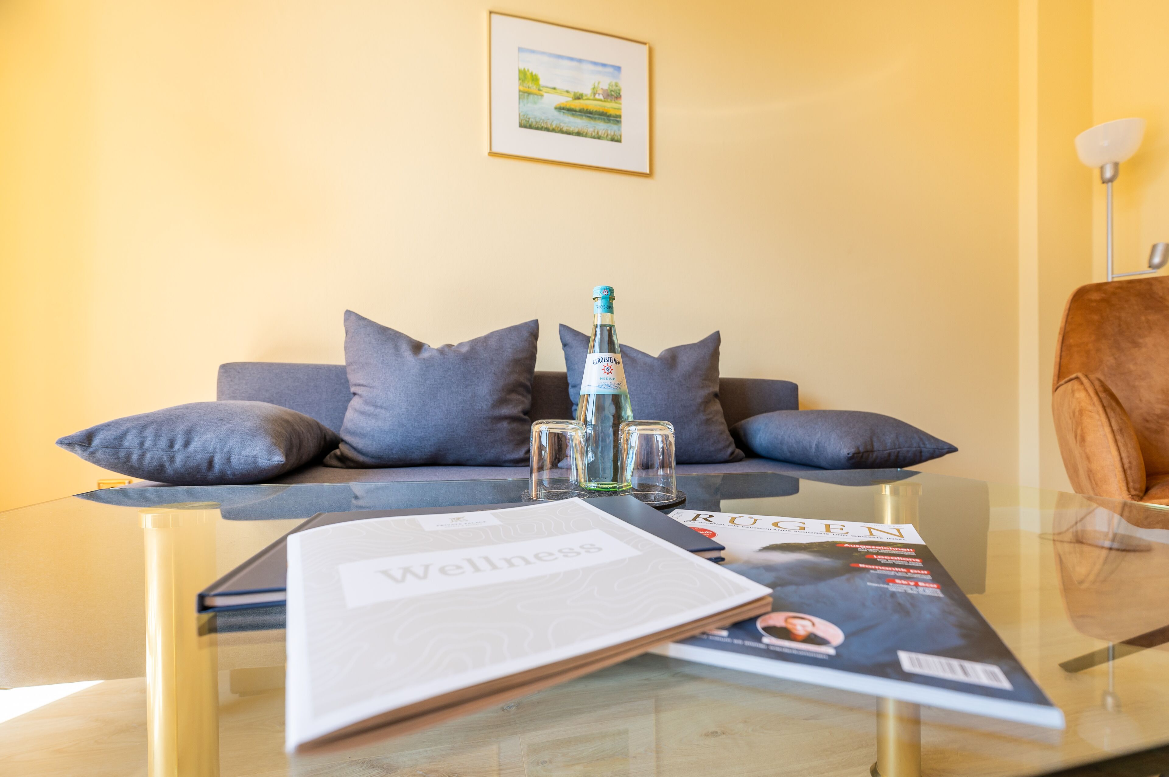 A living room with a sofa, pillows, a glass coffee table, a bottle, and magazines.