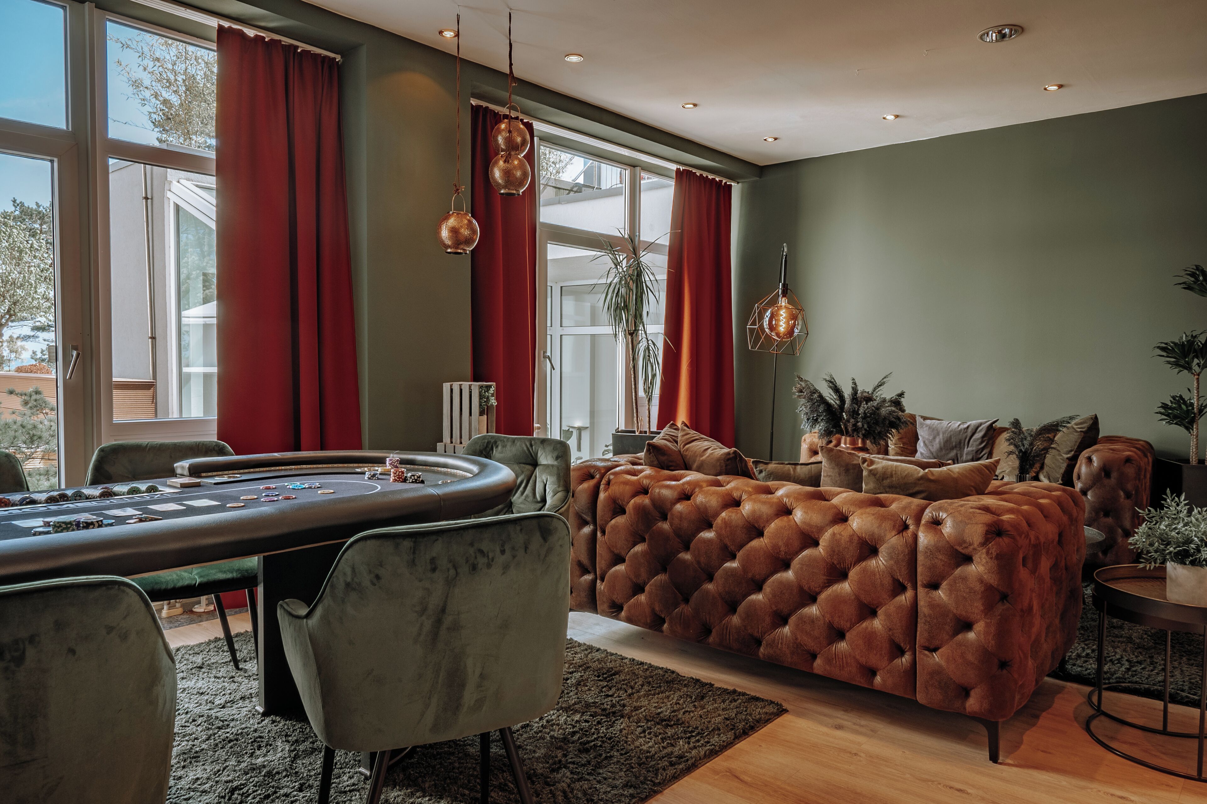 A modern living room with a large, tufted sofa, a poker table, and a view of the outdoors through large windows.