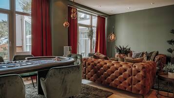 A modern living room with a large, tufted sofa, a poker table, and a view of the outdoors through large windows.