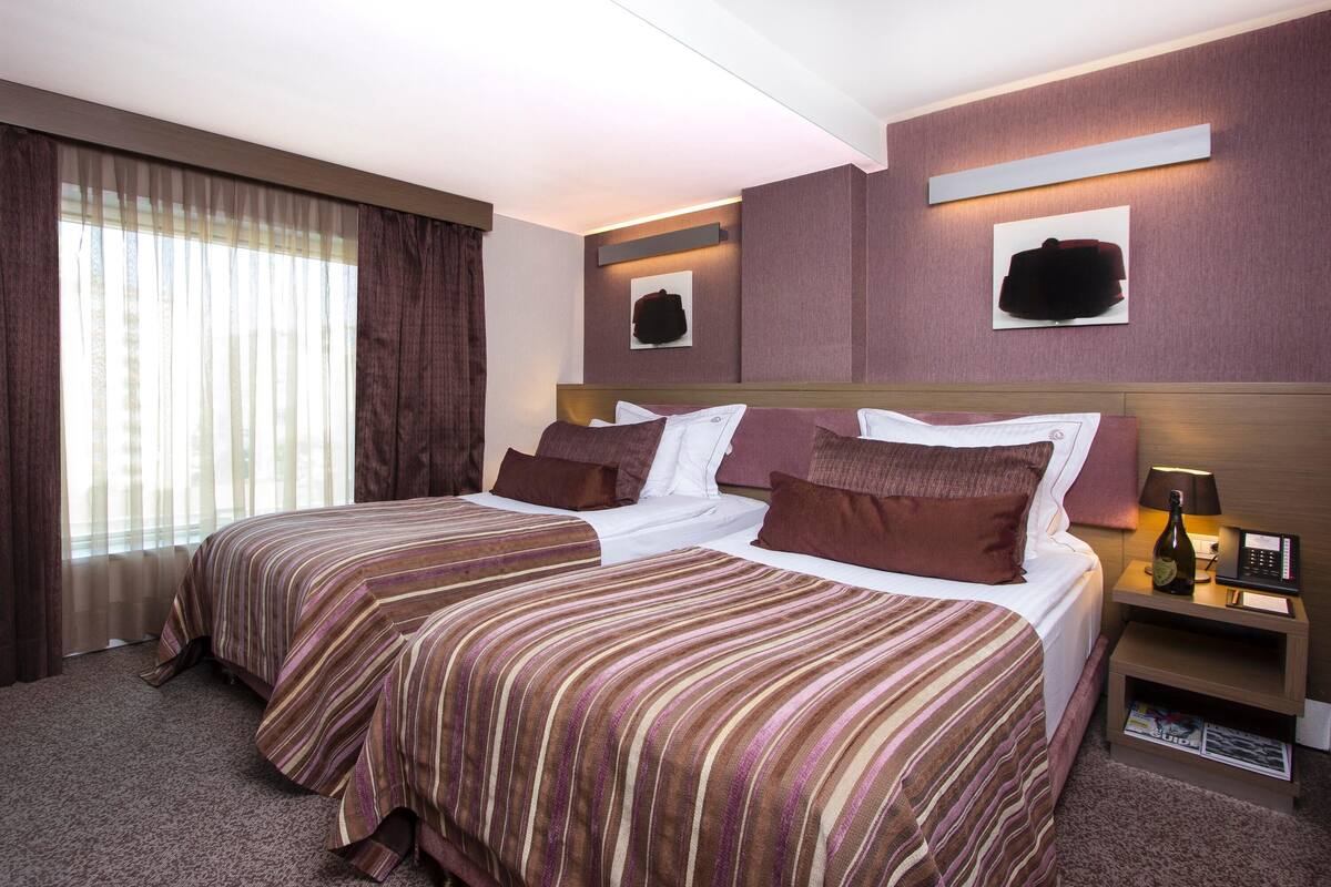 Quarto de hotel com duas camas, uma mesinha de cabeceira com abajur, uma garrafa de champanhe e obras de arte nas paredes.