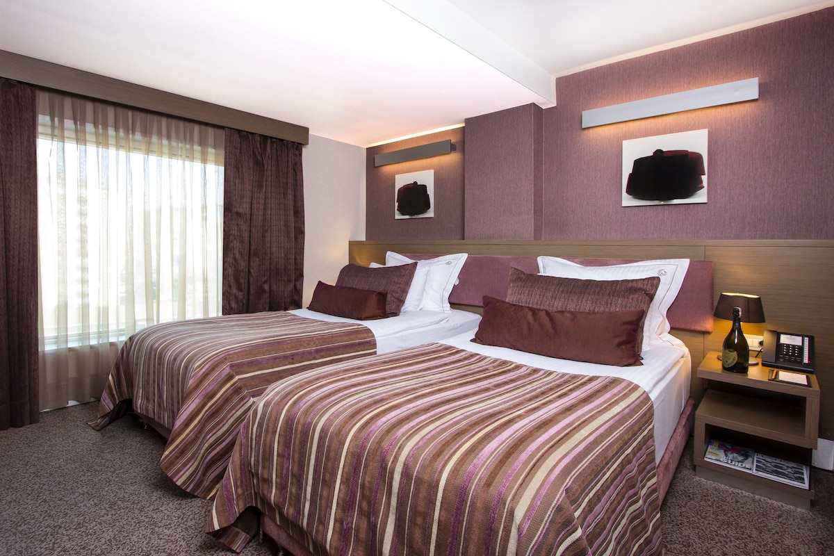 Une chambre d’hôtel avec deux lits, une table de chevet avec une lampe, une bouteille de champagne et des œuvres d’art accrochées aux murs.