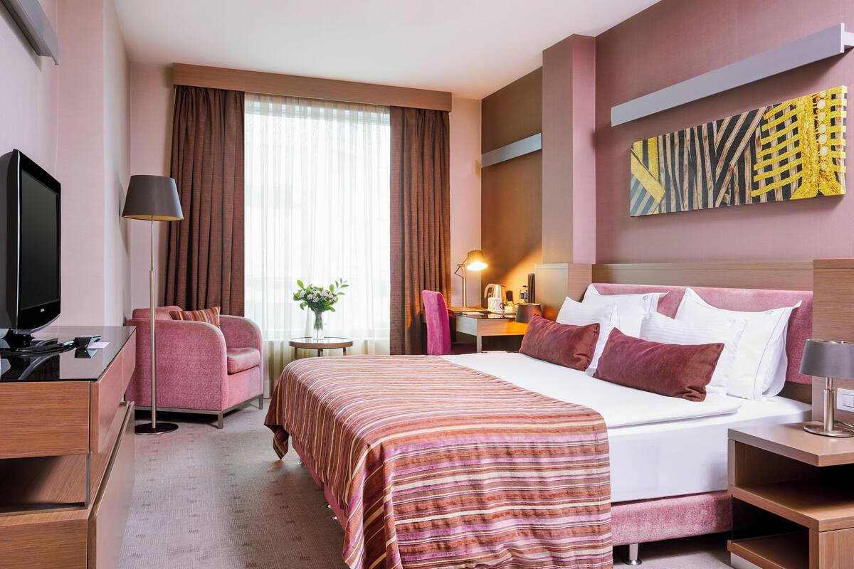 Quarto de hotel com cama, escrivaninha, cadeira, televisão e um quadro na parede.