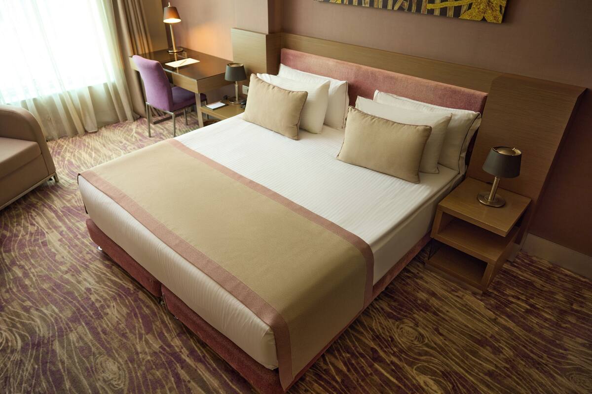 A hotel room with a large bed, a desk, a chair, and a nightstand with a lamp.