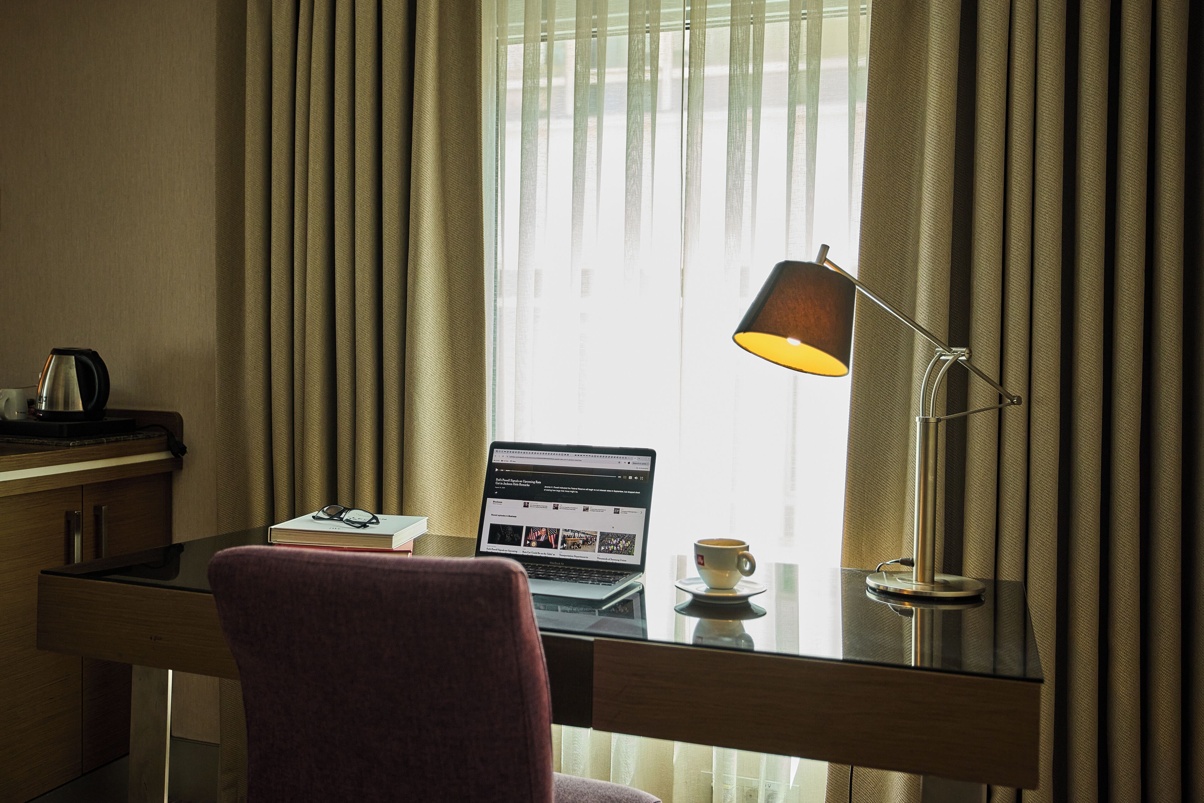 Une chambre d’hôtel avec un bureau, un ordinateur portable, une tasse de café et des rideaux.