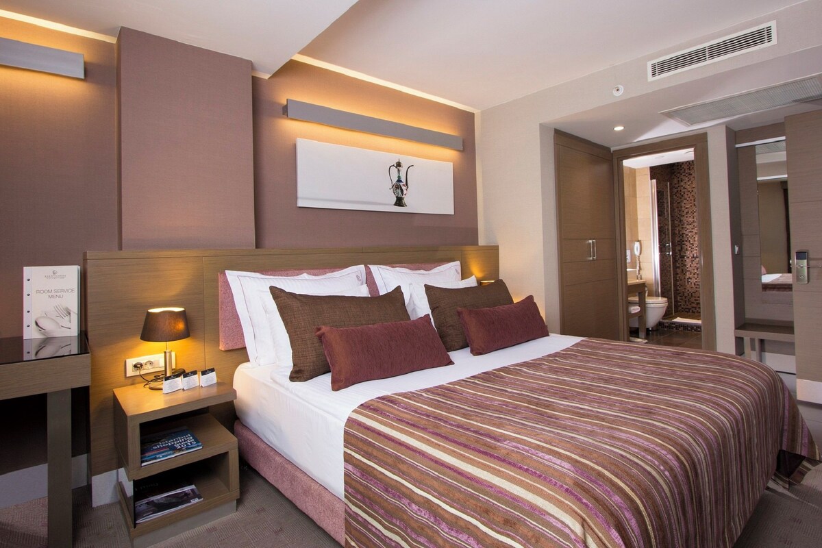 Une chambre d’hôtel avec un grand lit, une table de chevet avec une lampe, un tableau encadré au mur et une salle de bain visible à travers une porte ouverte.
