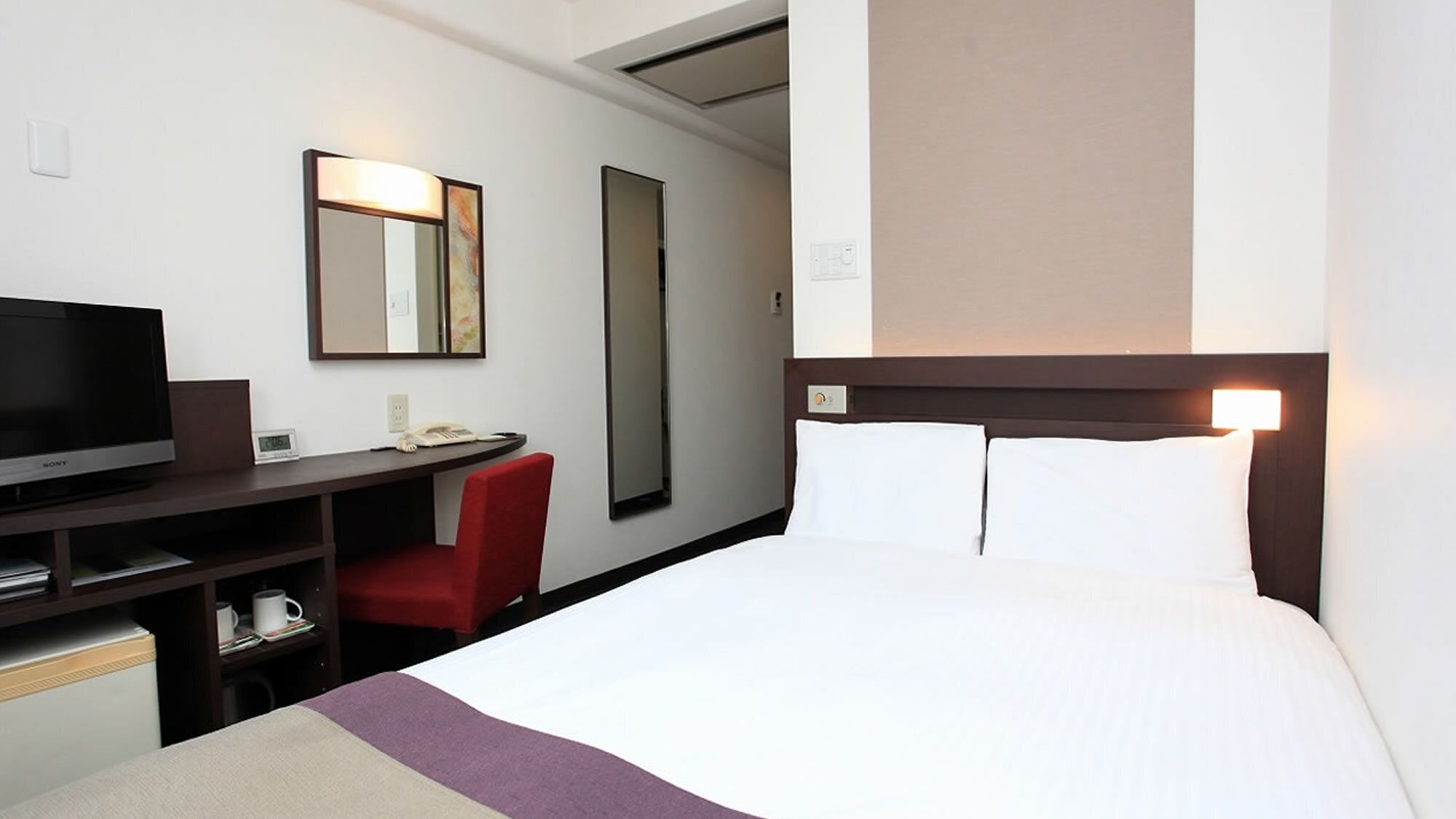 A hotel room with a bed, a desk with a TV, a chair, and a mirror.