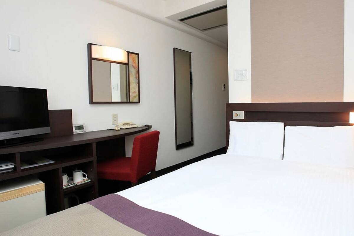 A hotel room with a bed, a desk with a TV, a chair, and a mirror.