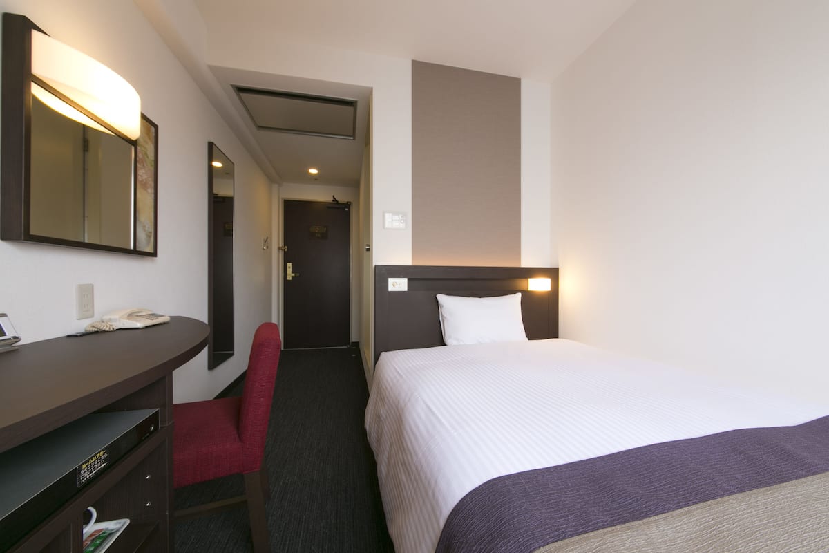 A hotel room with a bed, a desk, a chair, a mirror, and a nightstand.