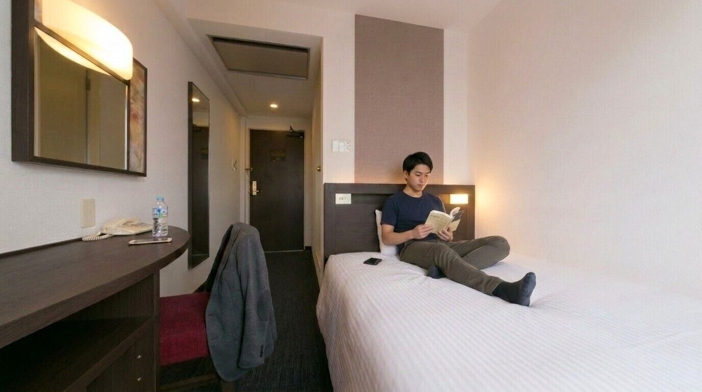 Una camera d'albergo con un letto, un comodino con un telefono e una bottiglia, uno specchio e una porta che dà su un'altra stanza.