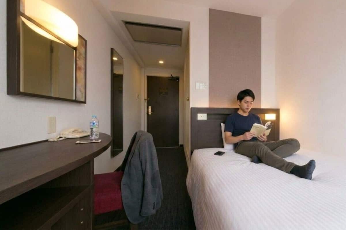 Una camera d'albergo con un letto, un comodino con un telefono e una bottiglia, uno specchio e una porta che dà su un'altra stanza.