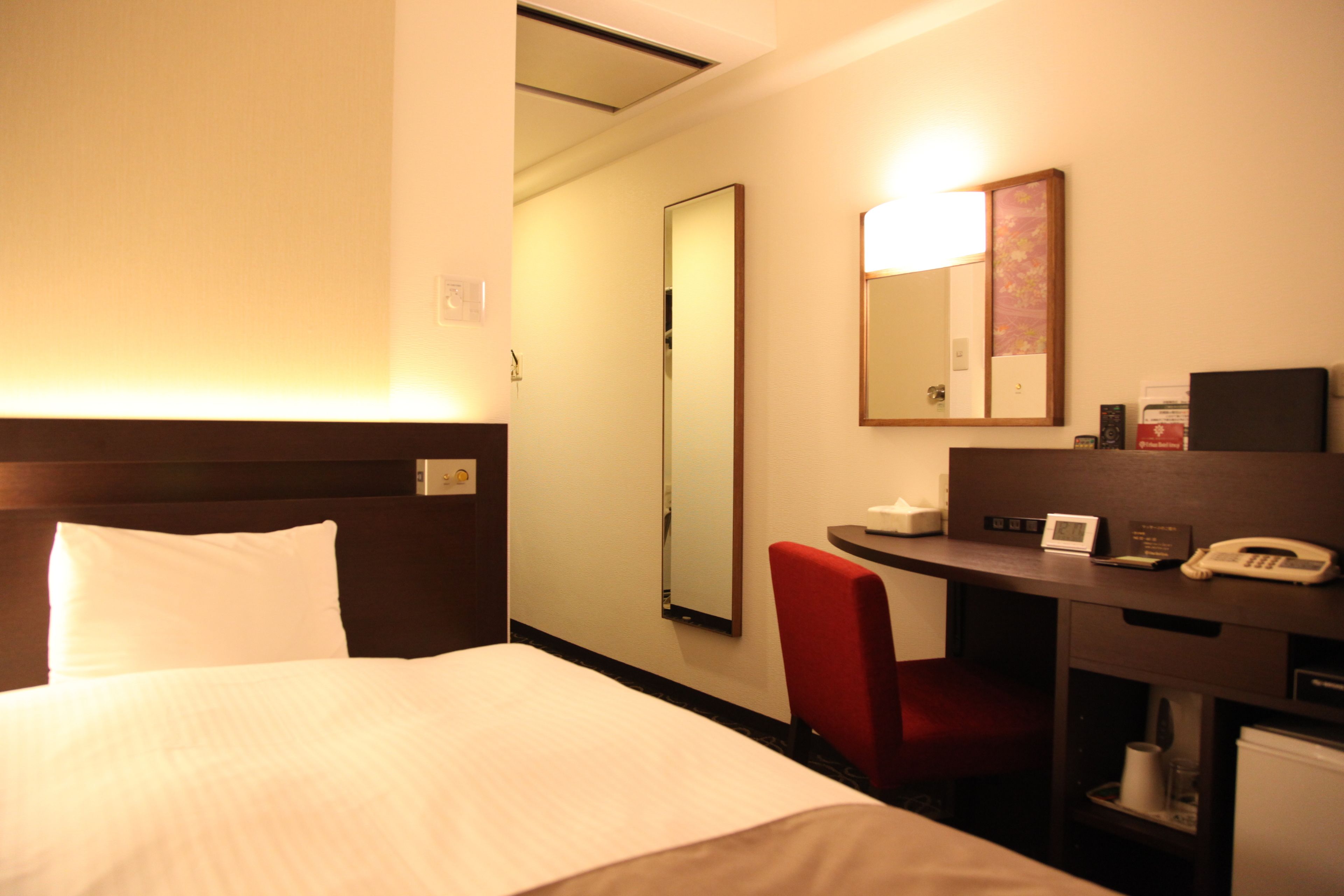 A hotel room with a bed, a desk with a telephone, a chair, a mirror, and a wall-mounted light.
