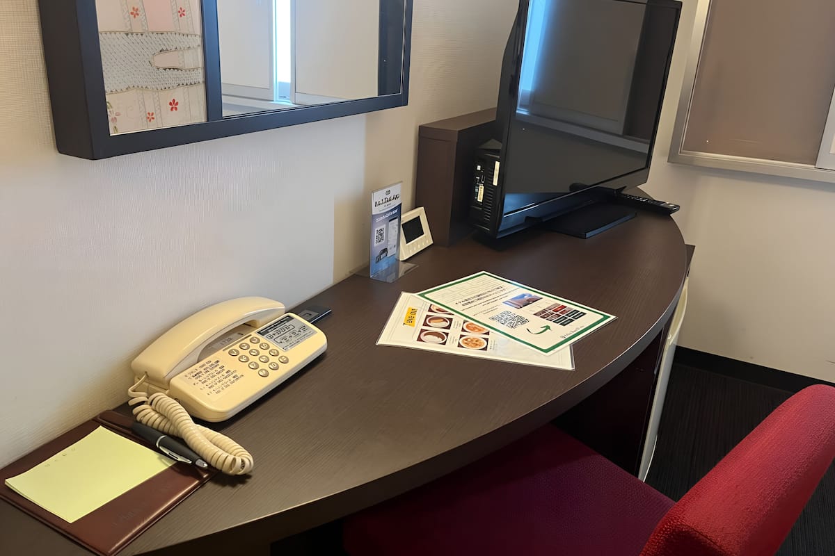 A hotel room desk with a telephone, a computer monitor, a desk lamp, a notepad, and a pen.