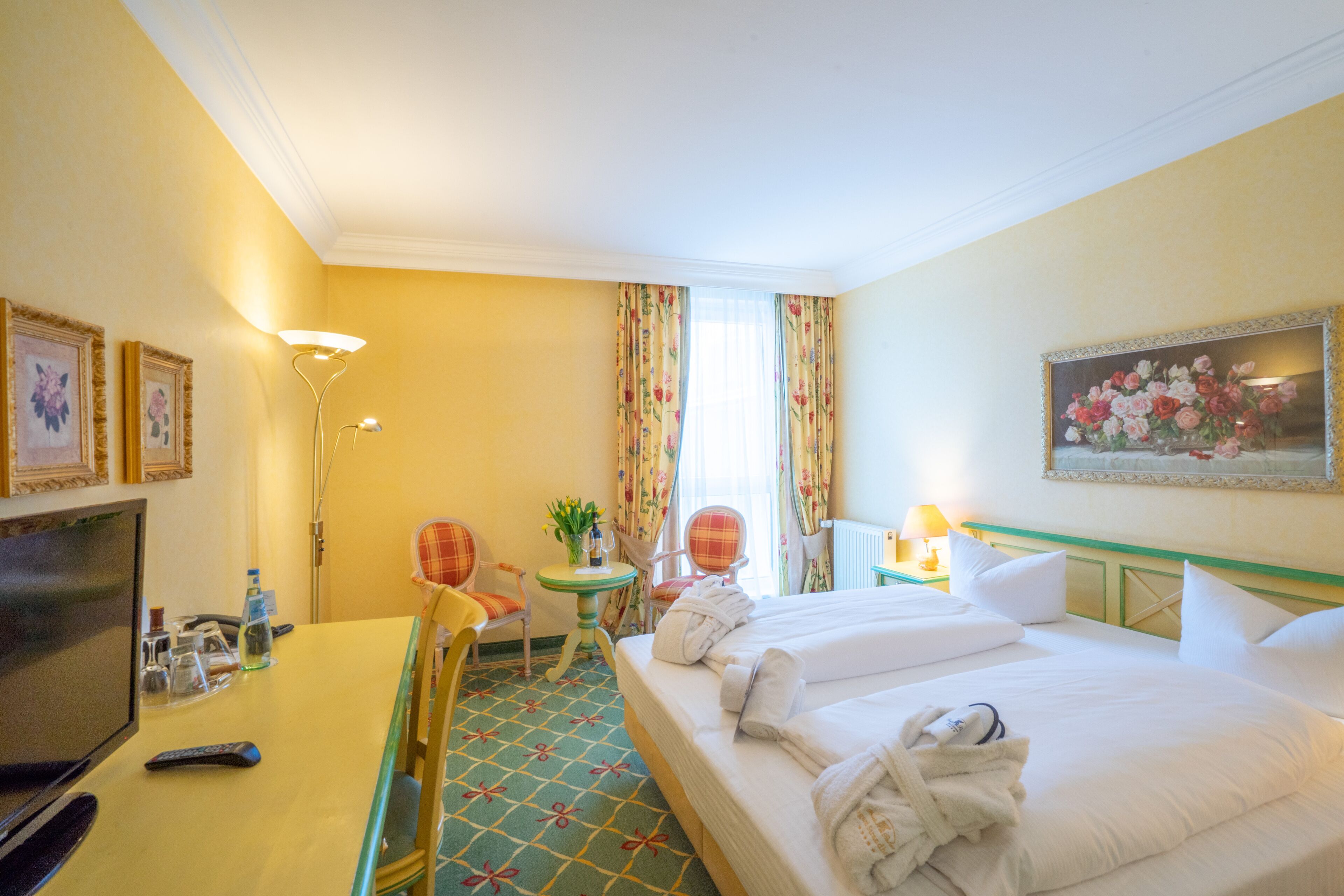 A hotel room with a bed, a desk with a TV, a chair, a small table with a lamp, and a painting on the wall.