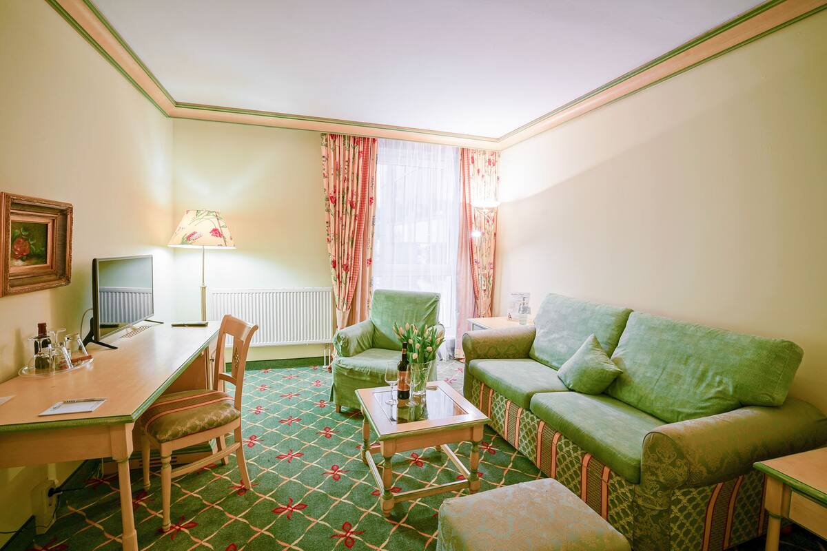 Une pièce avec un tapis à motifs verts, un canapé vert, un fauteuil vert, une petite table avec une lampe, un bureau avec un ordinateur et un tableau encadré accroché au mur.