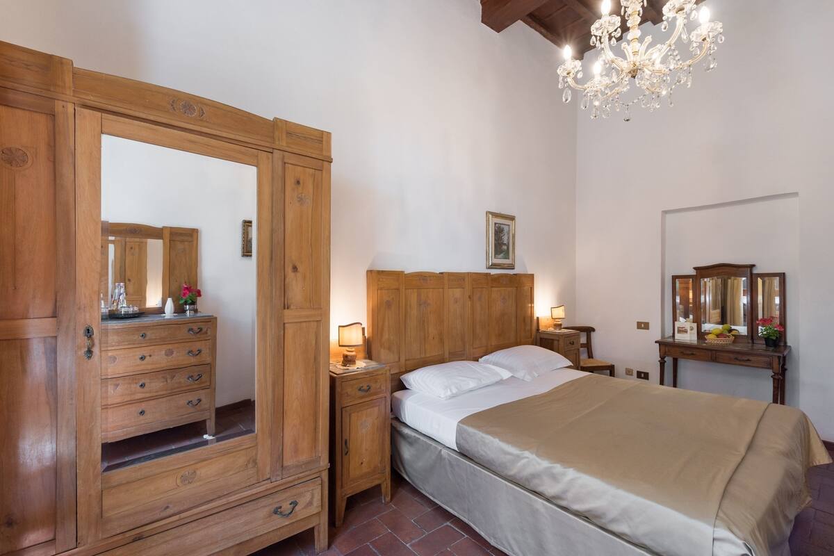 A bedroom with a wooden wardrobe, a large mirror, a bed with white linens, a wooden nightstand, and a chandelier.