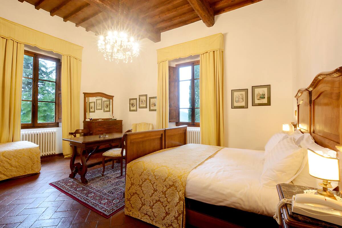 A hotel room with a large bed, a wooden dresser, a desk, and a chair. There are two windows with curtains, a radiator, and a chandelier.