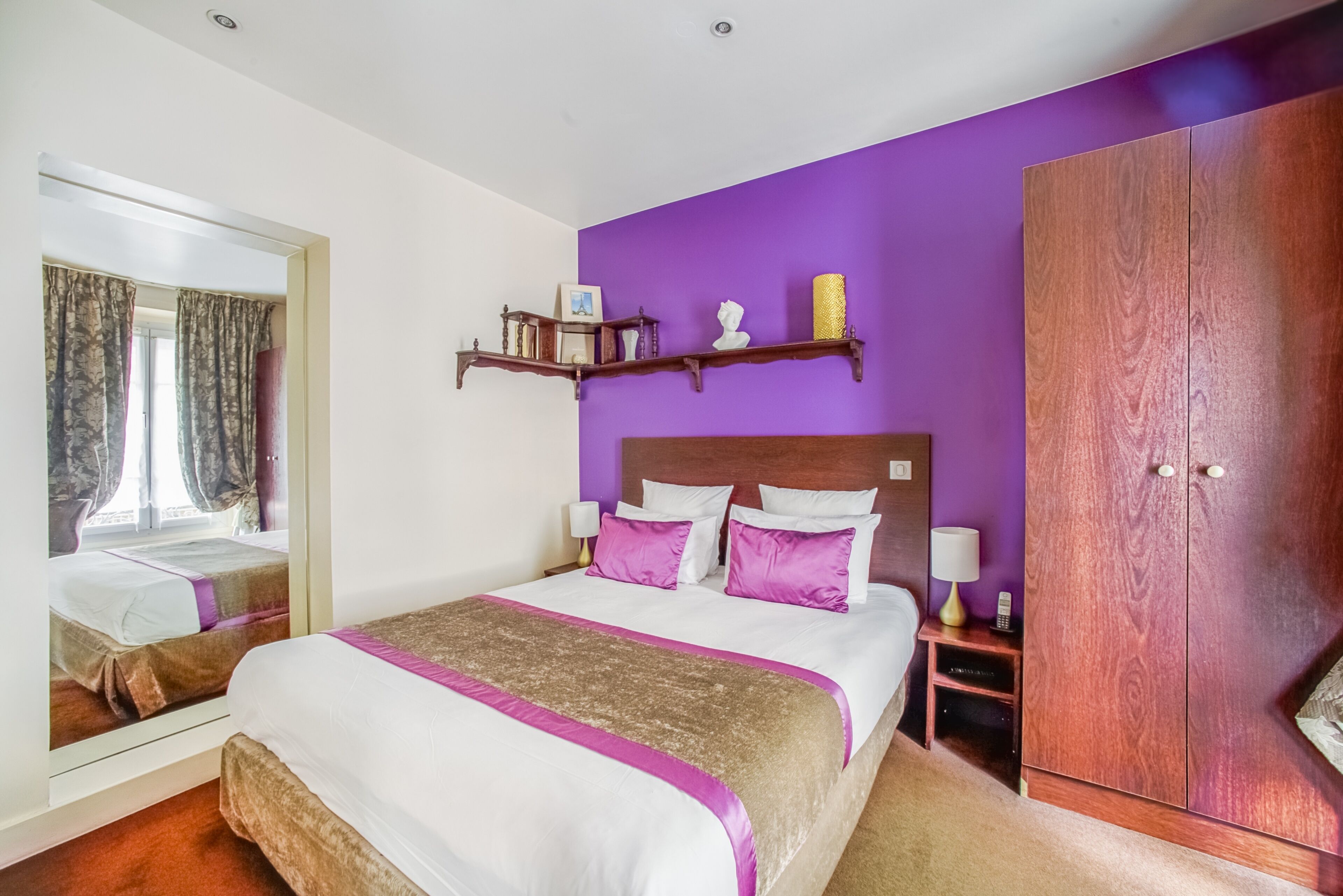Un dormitorio con una cama grande, paredes moradas, un armario de madera y un espejo.
