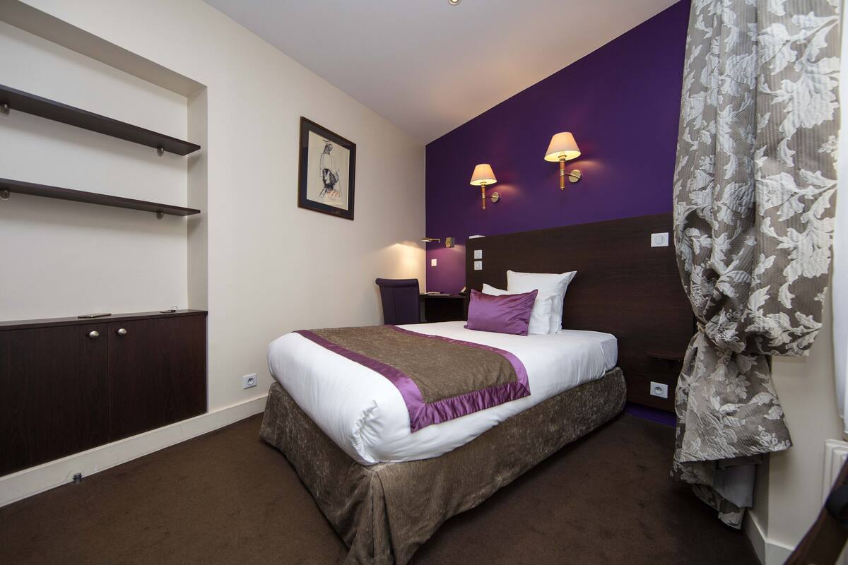 A neatly arranged hotel room with a large bed, dark wood paneling, purple accent walls, and two wall-mounted lamps.