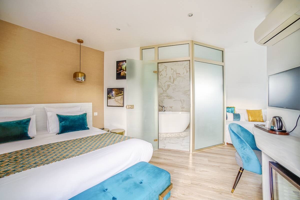 A modern hotel room with a large bed, a desk with a chair, a shower area, and a TV.