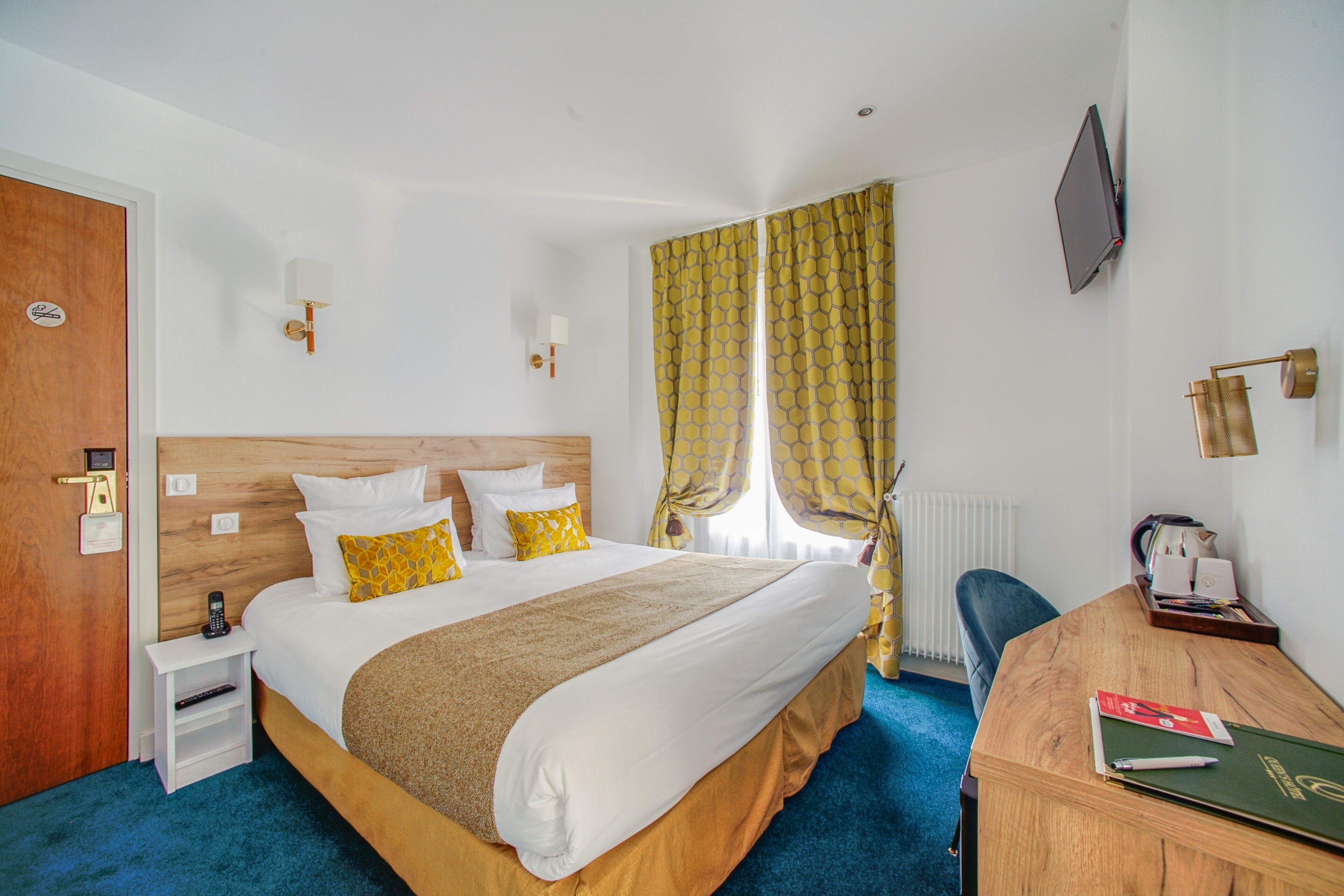 Habitación de hotel con una cama grande, un cabecero de madera, un escritorio con una silla, un televisor montado en la pared y una ventana con cortinas.