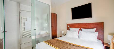 Habitación de hotel con cama, televisor montado en la pared, cabina de ducha de vidrio y armario de madera.