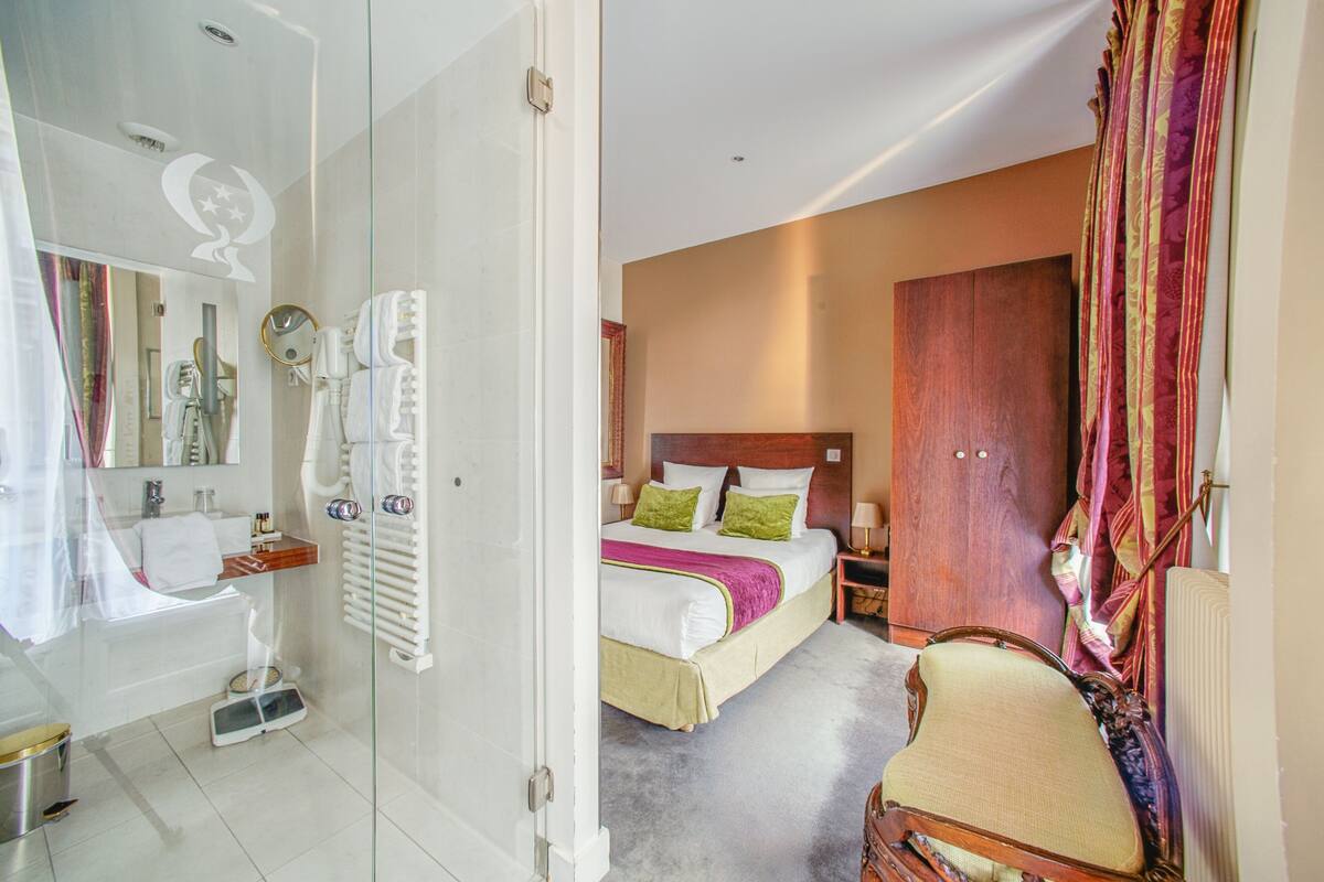 A hotel room with a glass shower enclosure, a bed with green pillows, a wooden headboard, and a large mirror.