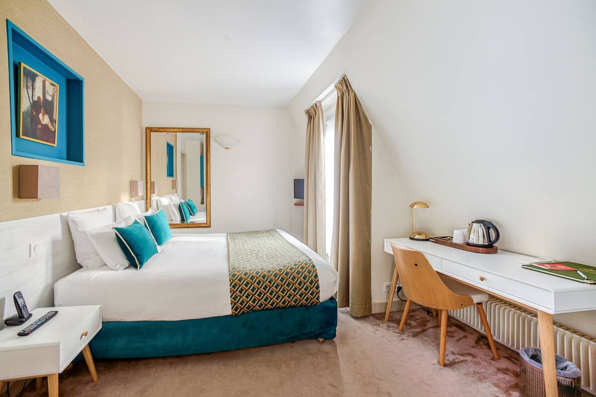 A hotel room with a bed, bedside table, desk, chair, and a window with curtains.