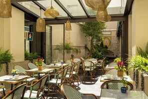 An indoor dining area with wicker furniture, glass tables, and hanging pendant lights. - Hôtel Muguet (Paris)
