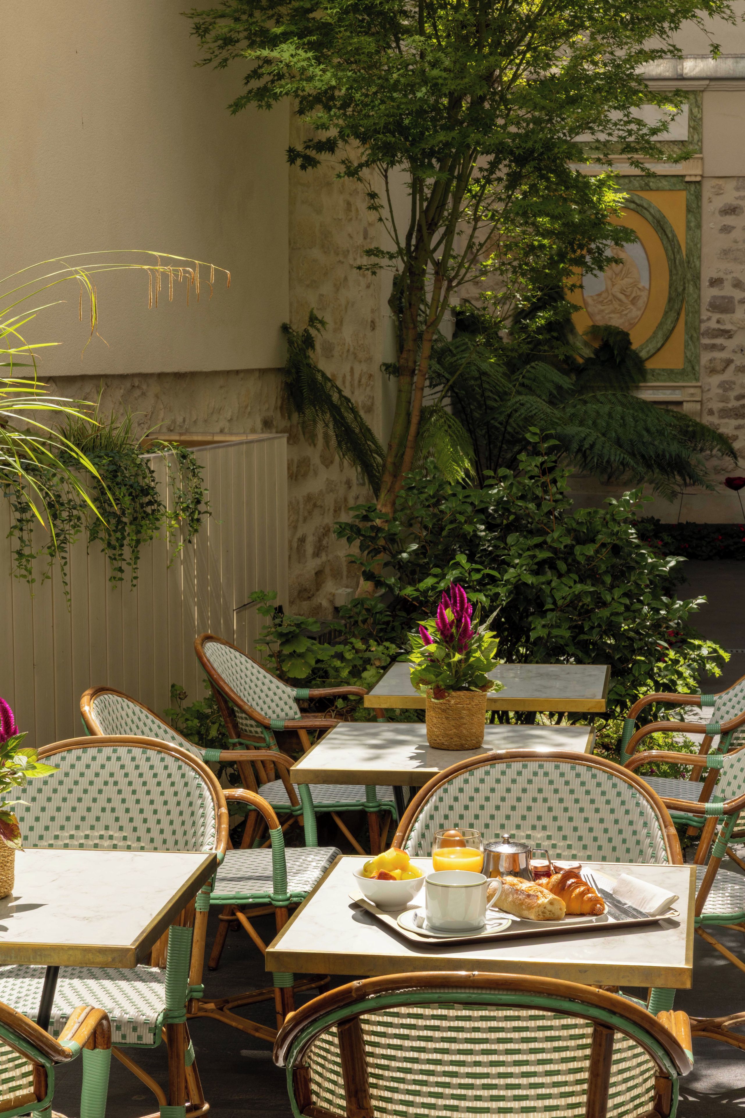 Un espace extérieur aménagé avec des chaises et des tables en osier, entouré de verdure et bordé d’un mur en pierre.