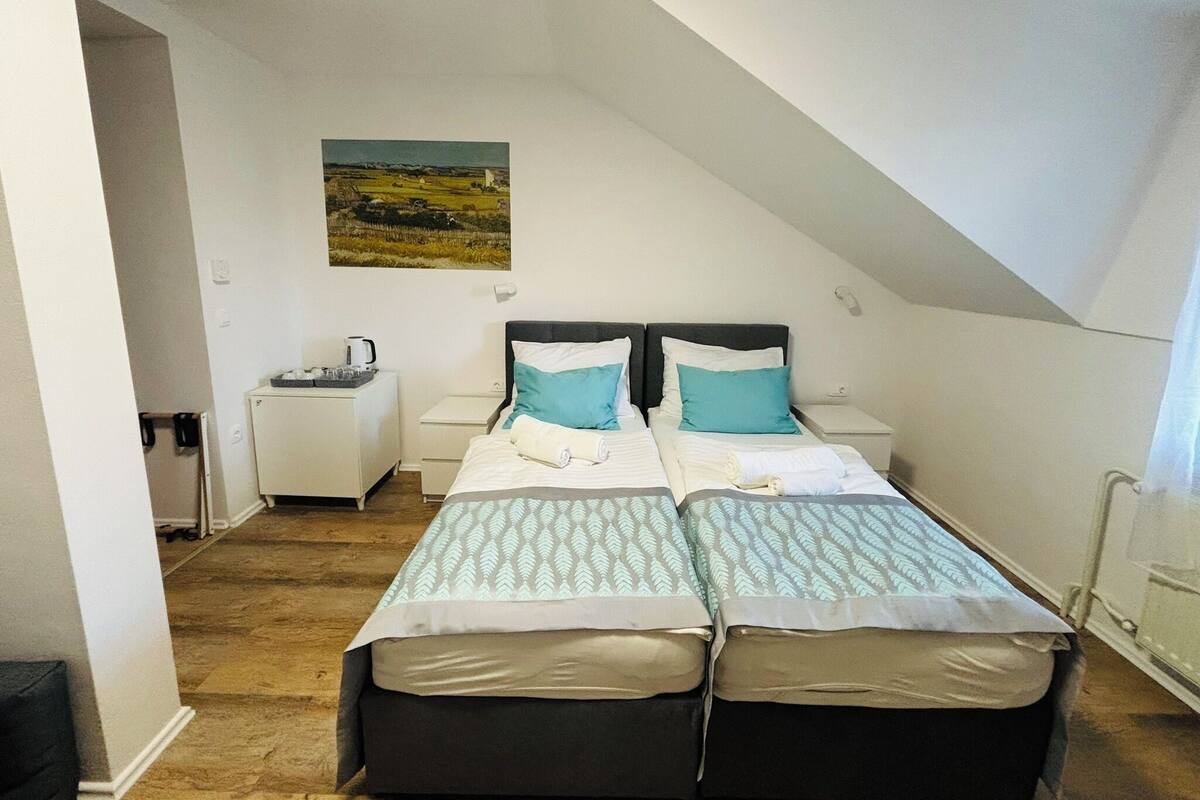 A small, well-lit room with two beds, a nightstand, a small table with a kettle, and a painting on the wall.