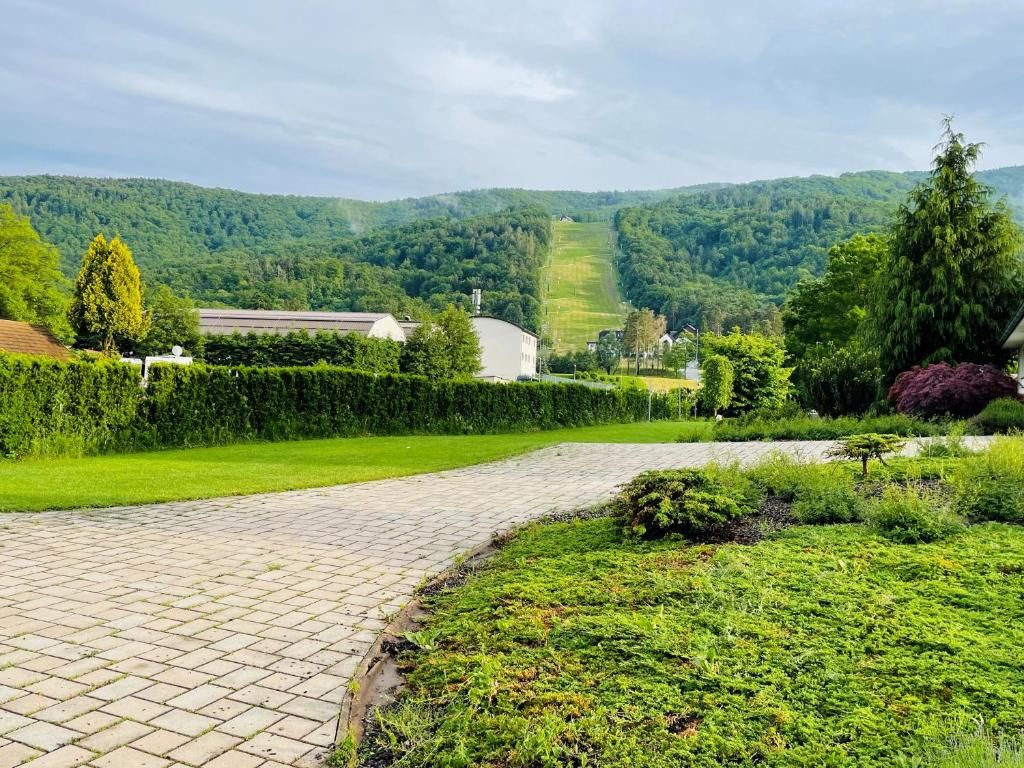 Un camino pavimentado que atraviesa un jardín ajardinado, con vistas a una colina y edificios al fondo.