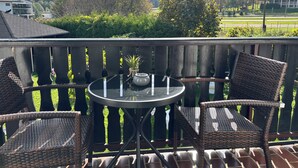 Een balkon met rieten stoelen en een tafel, met uitzicht op een groen landschap met bomen en een gebouw op de achtergrond.