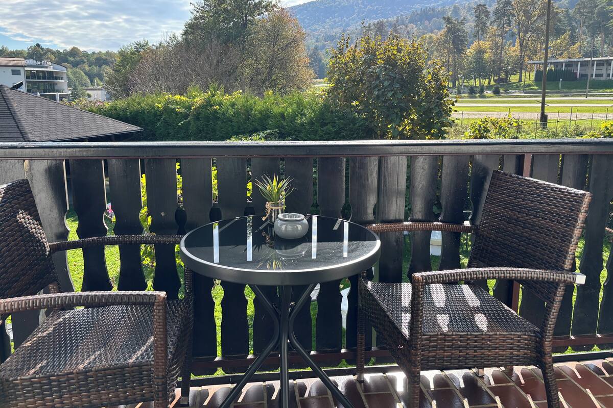 A balcony with wicker chairs and a table, overlooking a green landscape with trees and a building in the background.