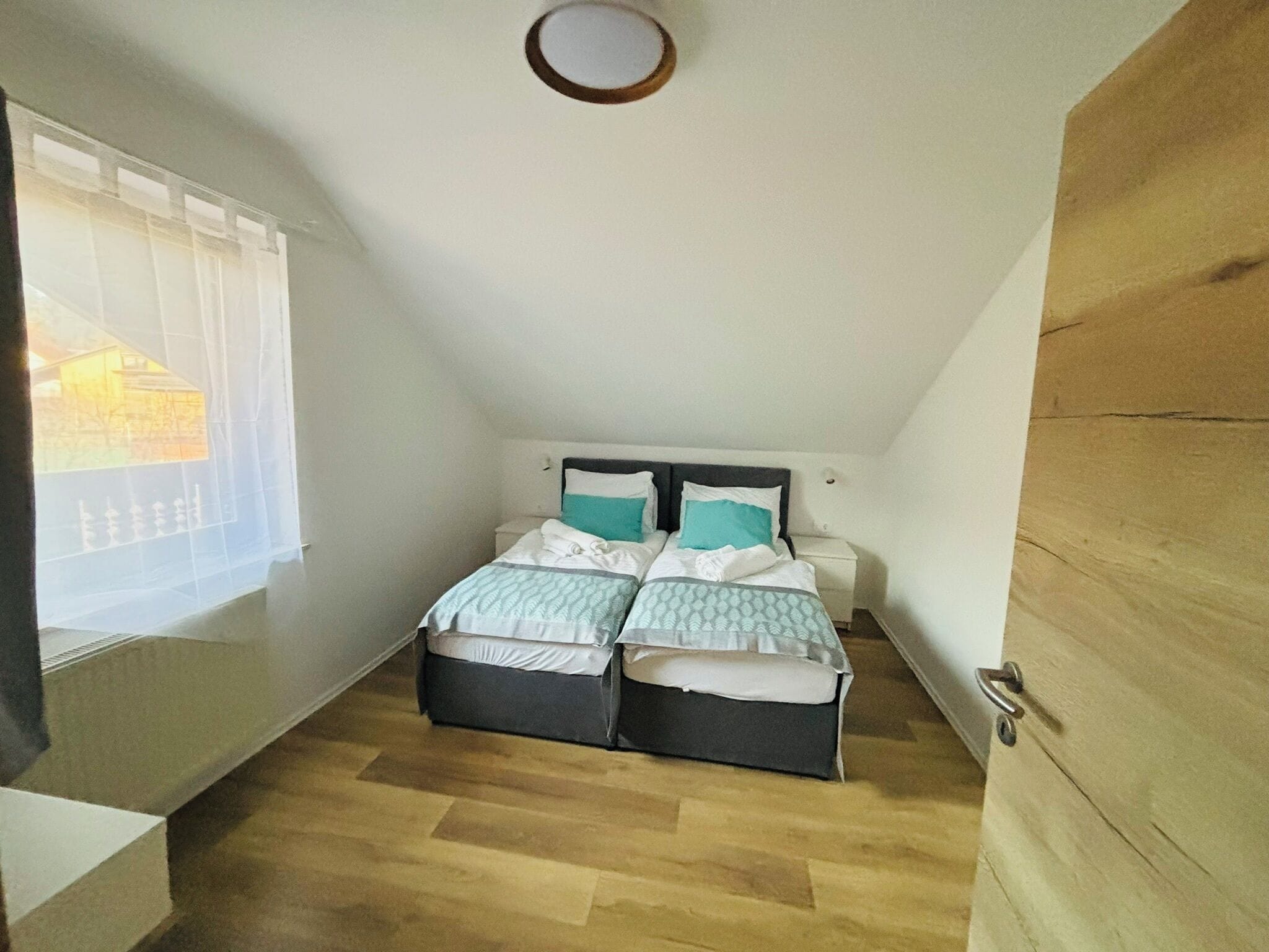 Um pequeno apartamento de um cômodo com teto inclinado, uma cama com duas almofadas, mesa de cabeceira e janela com cortinas leves.