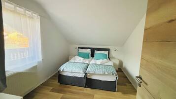 Um pequeno apartamento de um cômodo com teto inclinado, uma cama com duas almofadas, mesa de cabeceira e janela com cortinas leves.