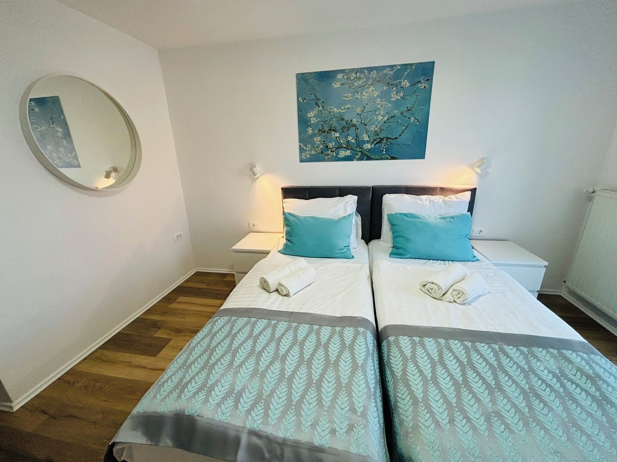 A double room with two beds, a round mirror, a painting, and a radiator.
