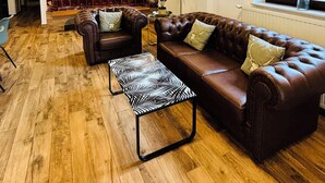 Een ruime woonkamer met een leren bank, een gestreept voetenbankje, en een houten vloer. Achtergrond is een keuken met een vast eiland en een eethoek met een tafel en stoelen.