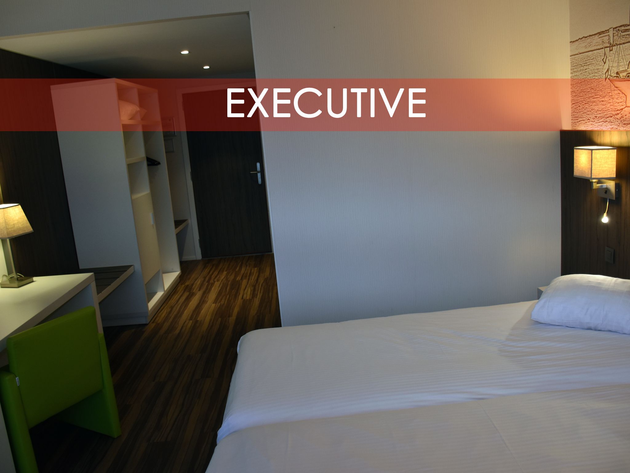 A hotel room with a bed, a desk, a chair, and a nightstand with a lamp.