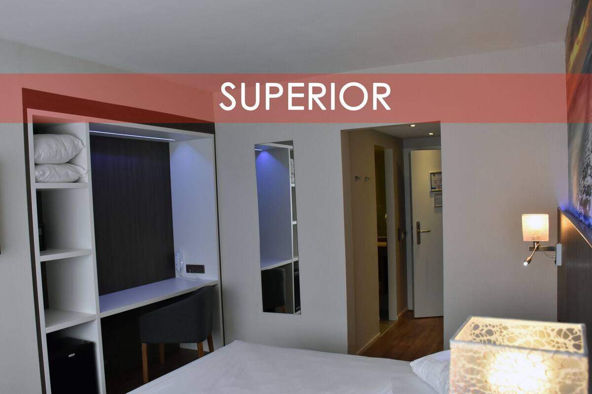 A hotel room with a bed, a wardrobe, a mirror, a door, and a wall-mounted lamp.