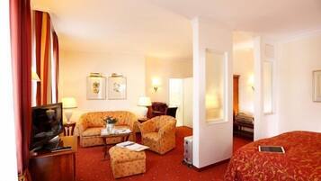 A hotel room with a red carpet, two armchairs, a sofa, a TV, a bedside table, a lamp, and a bed with a red cover.