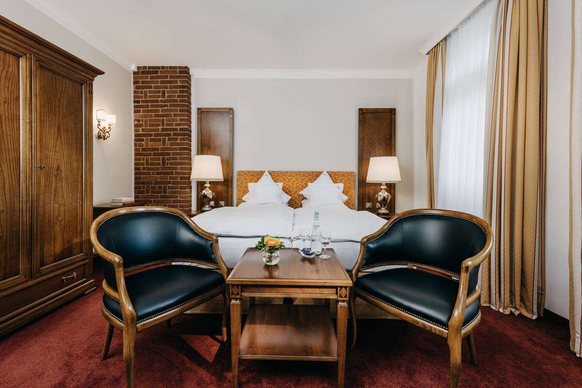 A hotel room with a large bed, two chairs, a wooden table, a brick accent wall, and a wardrobe.