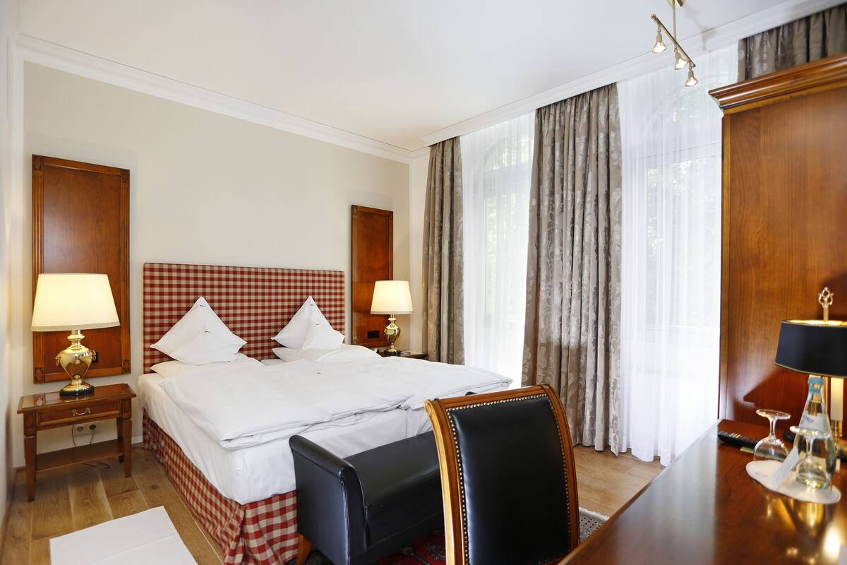 A hotel room with a large bed, two bedside tables with lamps, a wooden desk with a chair, and a large window with curtains.