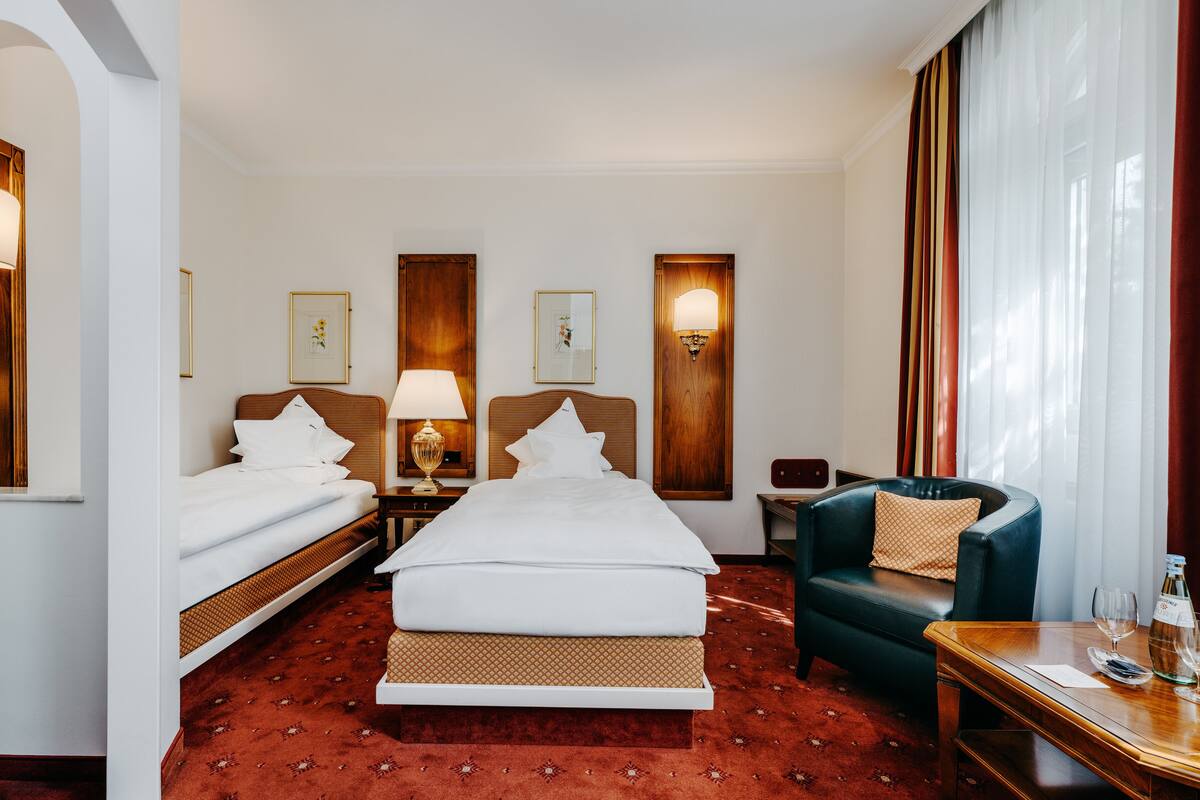 A hotel room with two beds, a sofa, a table with a bottle and glasses, and a window with curtains.