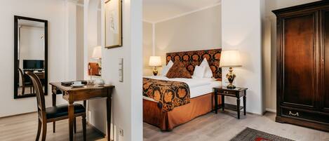 Habitación de hotel con un escritorio de madera, una cama con cabecera estampada, un espejo grande y un armario de madera oscura.