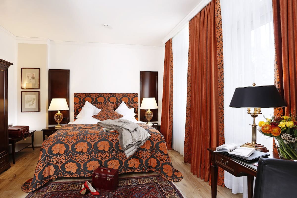 A hotel room with a large bed, two bedside tables with lamps, a desk with a lamp, a chair, a suitcase, and a vase with flowers.