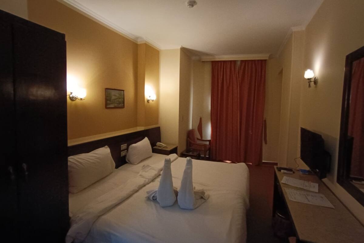 Quarto de hotel com duas camas, uma escrivaninha, uma cadeira, uma televisão e uma janela com cortinas.